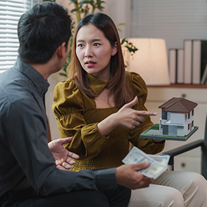 Young asian couple arguing about house investment and mortgage payment, woman pointing at house model while man holding us dollar banknotes, real estate and relationship concept