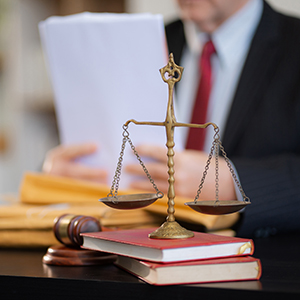 A gavel and scales of justice resting on red law books, with a blurred lawyer in the background.